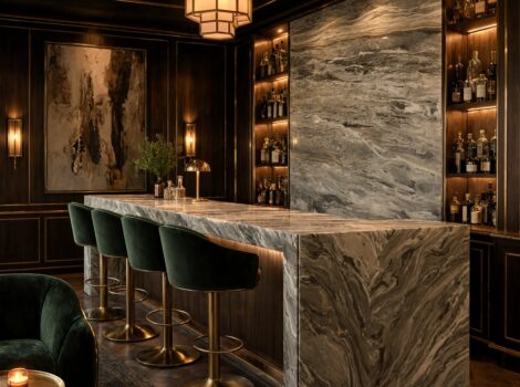 Fantasy brown marble bar countertop with waterfall edge and full back wall fantasy brown marble slab panel in a luxury home bar, green velvet bar stools with brass bases, dark wood paneling with gold trim and backlit bottle shelving
