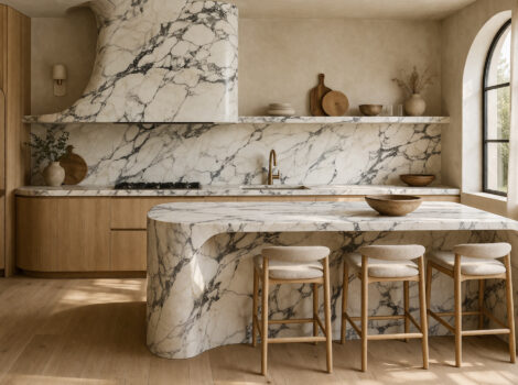 Monolithic statuario marble kitchen island with waterfall edge and bold gray veining, matching statuario marble slab backsplash and range hood cladding, paired with natural oak cabinetry and brass fixtures in an organic modern kitchen