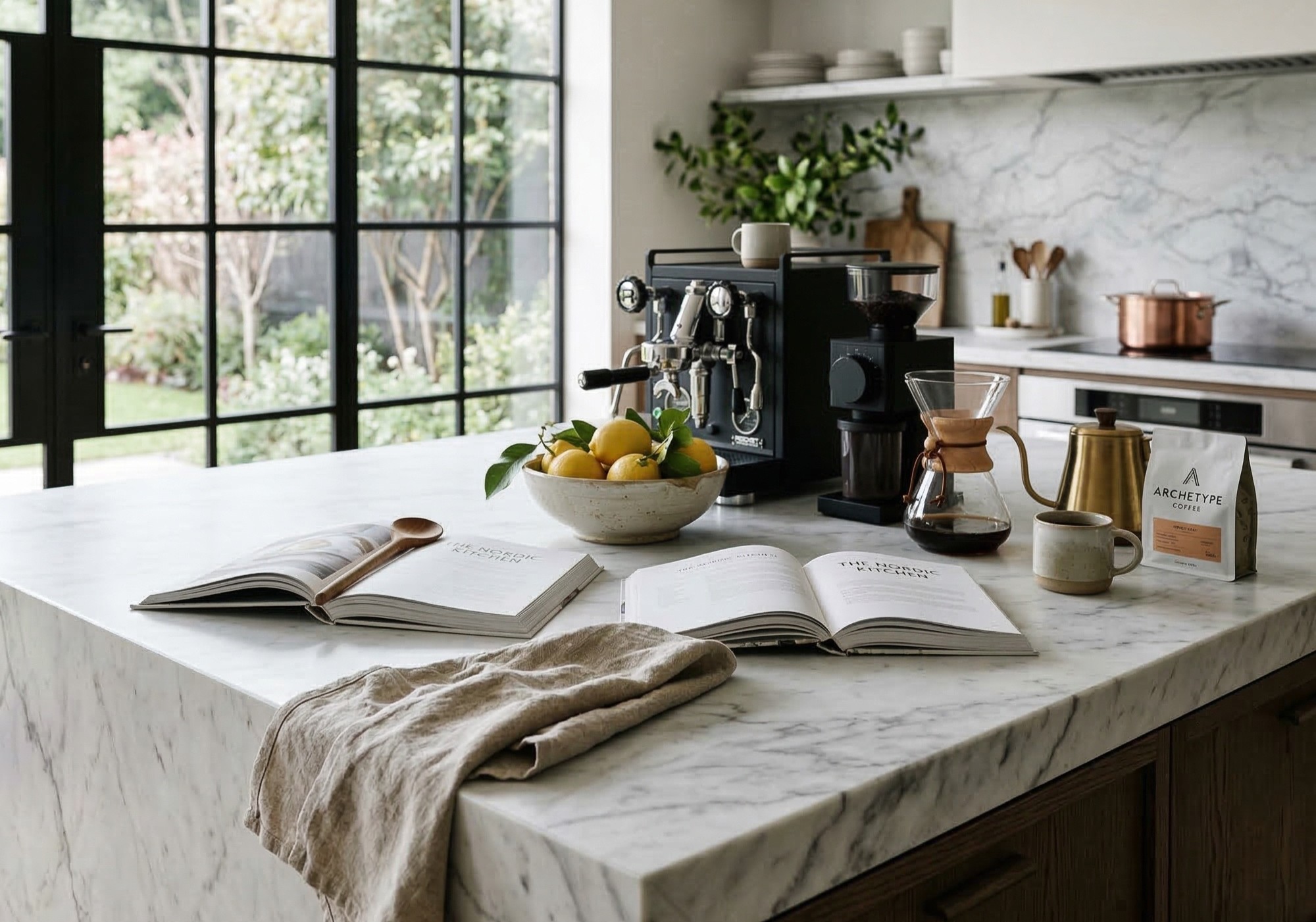 White carrara marble kitchen island countertop with subtle cool gray veining styled with open cookbooks, citrus bowl, professional espresso machine and Chemex pour over, carrara marble slab backsplash visible in background, black steel frame garden doors