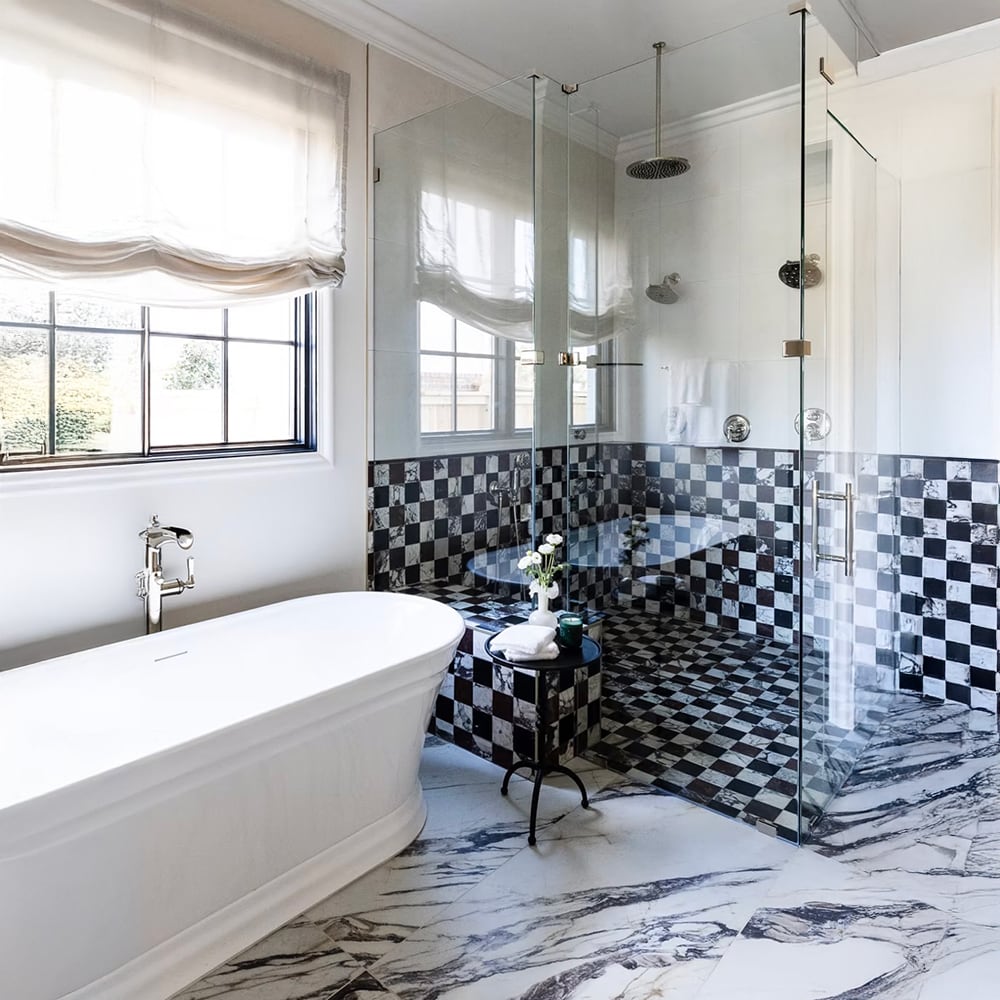 Calacatta viola marble mosaic tile in checkerboard pattern covering full shower wall and floor in black and white veined marble, freestanding white soaking tub with chrome fixtures, roman shade window with garden view in a luxury bathroom